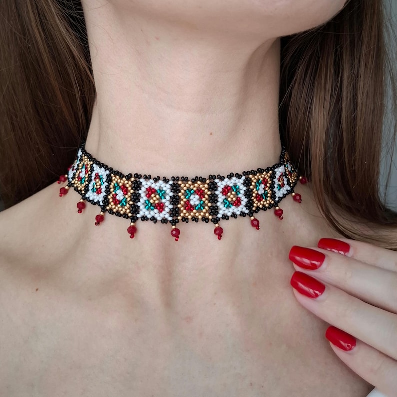 K&ouml;nnte beinhalten: Ein Perlen-Choker-Halskette mit schwarzer Basis, mit einem geometrischen Muster in Wei&szlig;, Rot, Gold und Gr&uuml;n. Kleine rote Perlen h&auml;ngen von der Unterkante. Die Halskette wird um den Hals getragen.