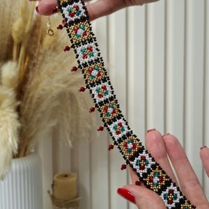 K&ouml;nnte beinhalten: Eine Halskette mit Perlen im Choker-Stil mit einem geometrischen Muster in Rot, Wei&szlig;, Schwarz, Gold und T&uuml;rkis. Die Halskette hat kleine rote Perlen, die am unteren Rand h&auml;ngen. Der Verschluss ist goldfarben.