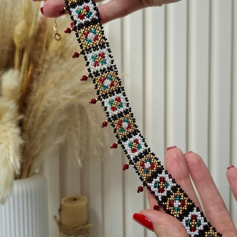 K&ouml;nnte beinhalten: Eine Halskette mit Perlen im Choker-Stil mit einem geometrischen Muster in Rot, Wei&szlig;, Schwarz, Gold und T&uuml;rkis. Die Halskette hat kleine rote Perlen, die am unteren Rand h&auml;ngen. Der Verschluss ist goldfarben.
