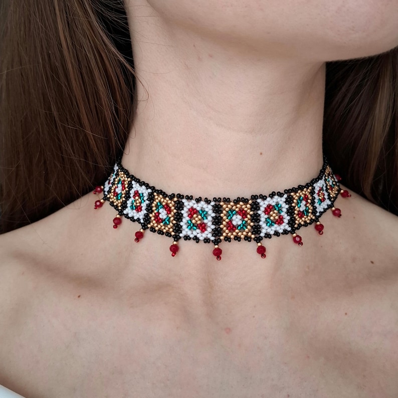 K&ouml;nnte beinhalten: Ein Perlen-Choker-Halskette mit schwarzer Basis, mit einem sich wiederholenden Muster aus wei&szlig;en, roten, gr&uuml;nen und goldenen Perlen. Kleine rote Perlen h&auml;ngen von der Unterkante des Chokers.