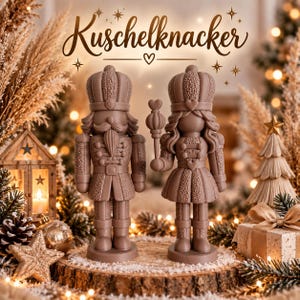 Könnte beinhalten: Zwei hellbraune Nussknacker-Figuren auf einem Holzsockel. Die linke Figur trägt einen Schnurrbart und eine Krone, die rechte hält ein Zepter mit Herzspitze. Darüber steht der Schriftzug 'Kuschelknacker' in einer dekorativen Schreibschrift.