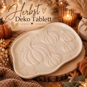 Puede incluir: Una bandeja decorativa de color blanco roto con forma orgánica irregular que presenta diseños en relieve de varias calabazas y hojas de arce. El texto "Herbst Deko Tablett" está impreso en color marrón oscuro sobre la bandeja.