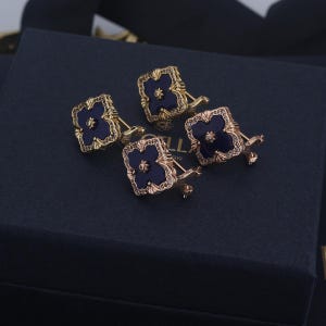 Peut inclure: Quatre boutons de manchette dorés avec un motif de trèfle bleu foncé. Chaque bouton de manchette a une forme carrée avec un motif floral et est orné de petits cristaux transparents. Les boutons de manchette sont présentés sur une surface bleu foncé.