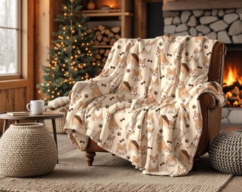 Könnte beinhalten: Beige Überwurfdecke mit einem Muster aus braunen und beigen Corgi-Hunden, Pfotenabdrücken und Knochen, drapiert über einem braunen Sessel. Im Hintergrund sind ein Weihnachtsbaum, ein Kamin und ein geflochtener Hocker zu sehen.