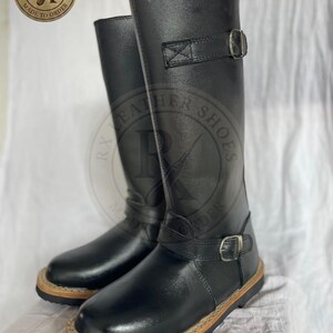 Könnte beinhalten: Ein Paar kniehohe Stiefel aus schwarzem Leder mit zwei verstellbaren Riemen und silberfarbenen Metallschnallen, einer am Knöchel und einer an der Wade. Die Stiefel haben eine abgerundete Zehenpartie und eine dicke schwarze Sohle mit sichtbarem beigen Rahmen.