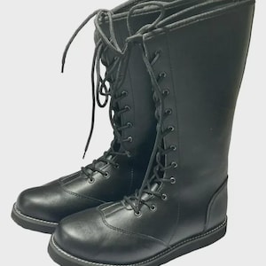 Puede incluir: Un par de botas negras hasta la rodilla con acabado liso, punteras redondeadas y cordones frontales completos con ojales negros y cordones a juego. Las botas tienen una suela plana negra gruesa con costuras blancas visibles a lo largo del borde.