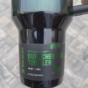 May include: A black and green Stanley Quencher H2.0 tumbler with a handle. The tumbler has the text "QUENCHER H2.0 TUMBLER" and "40 OZ | 1.18 L" printed on the side. The tumbler is designed for all-day hydration.