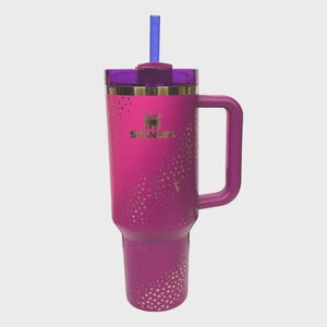 May include: A magenta Stanley tumbler with a handle, a purple lid, and a blue straw. The tumbler has a gold rim and a pattern of gold stars. The Stanley logo is visible on the front.