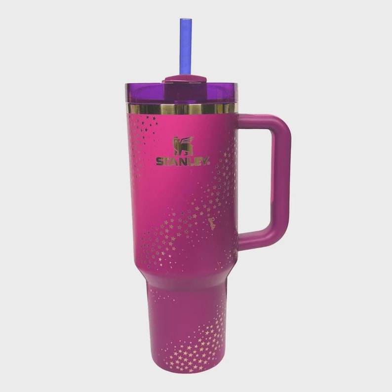 May include: A magenta Stanley tumbler with a handle, a purple lid, and a blue straw. The tumbler has a gold rim and a pattern of gold stars. The Stanley logo is visible on the front.