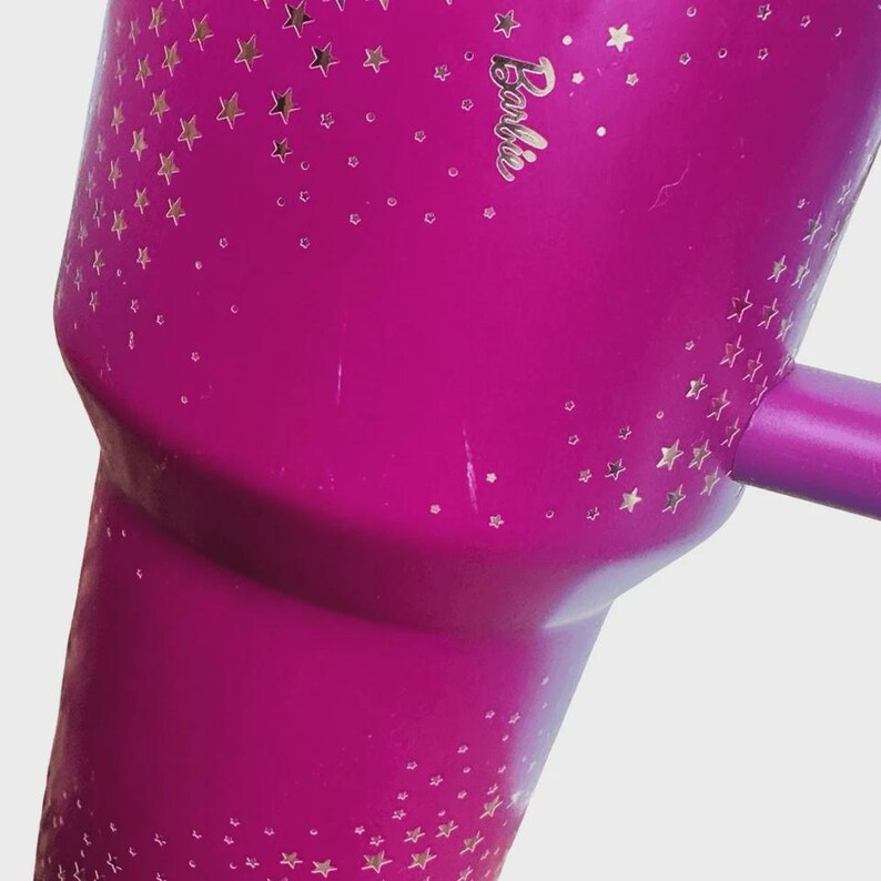 May include: A magenta tumbler with a handle, decorated with gold star patterns and the word "Barbie" in gold script. The tumbler has a smooth finish and a cylindrical shape, ideal for cold drinks.