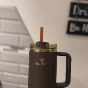 Puede incluir: Un termo de viaje Stanley marr&oacute;n con tapa y asa doradas. El termo tiene una pajita y el logotipo de Stanley en dorado. El termo est&aacute; dise&ntilde;ado para mantener las bebidas calientes o fr&iacute;as.