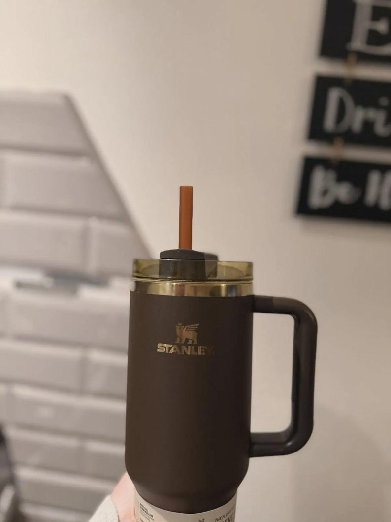 Puede incluir: Un termo de viaje Stanley marr&oacute;n con tapa y asa doradas. El termo tiene una pajita y el logotipo de Stanley en dorado. El termo est&aacute; dise&ntilde;ado para mantener las bebidas calientes o fr&iacute;as.