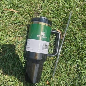 May include: A green and black Stanley Quencher Tumbler with a black handle and a silver straw, resting on a bed of green grass. The tumbler has the Stanley logo and the words "QUENCHER TUMBLER" printed on it.
