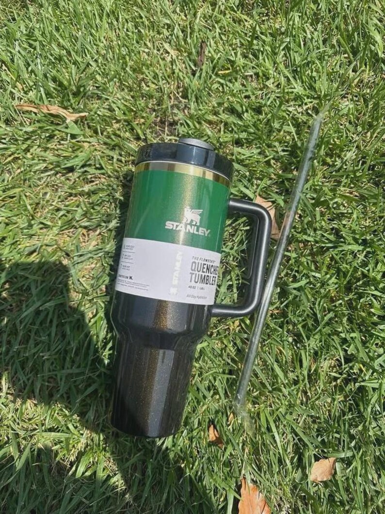 May include: A green and black Stanley Quencher Tumbler with a black handle and a silver straw, resting on a bed of green grass. The tumbler has the Stanley logo and the words "QUENCHER TUMBLER" printed on it.