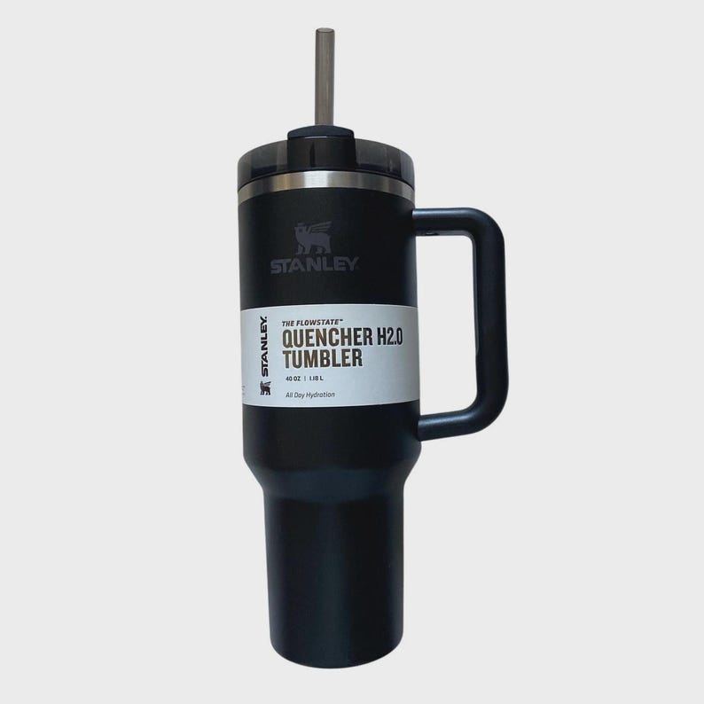 May include: Black Stanley Quencher H2.0 Tumbler with a handle and a straw. The tumbler has a black lid and a white label with the text "QUENCHER H2.0 TUMBLER" and the Stanley logo. It holds 40 oz (1.18 L).