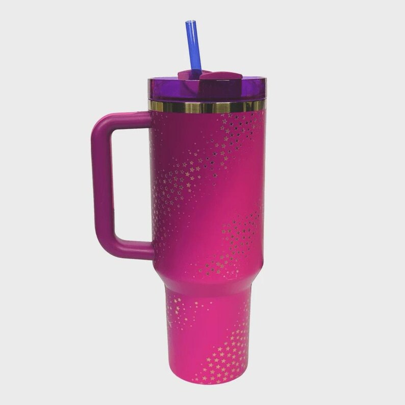 May include: A bright pink insulated travel mug with a handle, a purple lid, and a blue straw. The mug features a pattern of small, gold-coloured stars. A gold band accents the top of the mug.