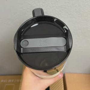 May include: A Stanley travel mug, viewed from above, featuring a black lid with a gray slider and the Stanley logo. The mug has a black handle and a cream-colored body. The image is taken indoors.