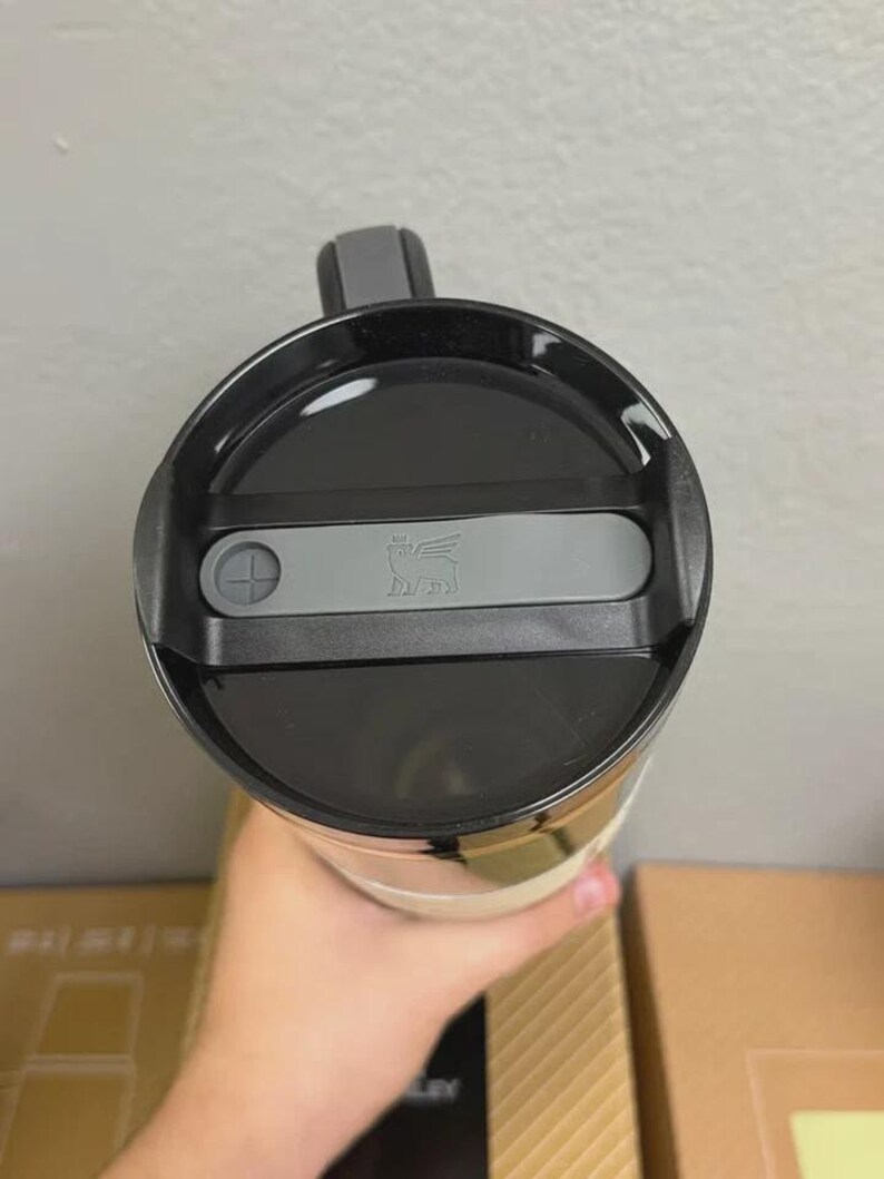 May include: A Stanley travel mug, viewed from above, featuring a black lid with a gray slider and the Stanley logo. The mug has a black handle and a cream-colored body. The image is taken indoors.