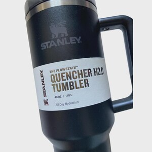 May include: Black Stanley Quencher H2.0 tumbler with a handle. The tumbler has the Stanley logo and text that reads "QUENCHER H2.0 TUMBLER" and "40 OZ | 1.18 L". The tumbler is designed for all-day hydration.
