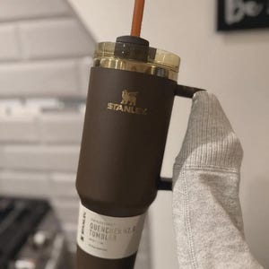 Puede incluir: Un vaso Stanley Quencher H2.0 marr&oacute;n con una tapa dorada y una pajita marr&oacute;n. El vaso tiene un asa y el logotipo de Stanley en dorado. El texto "QUENCHER H2.0 TUMBLER" es visible en la etiqueta.