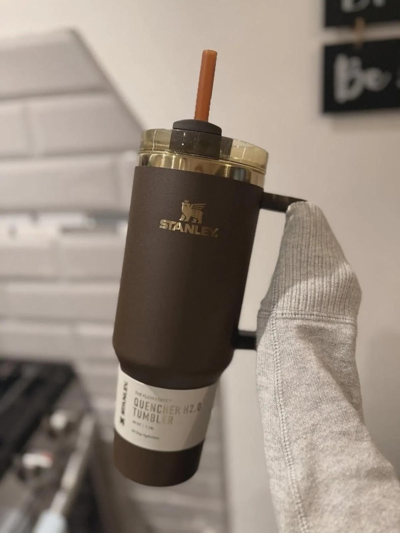 Puede incluir: Un vaso Stanley Quencher H2.0 marr&oacute;n con una tapa dorada y una pajita marr&oacute;n. El vaso tiene un asa y el logotipo de Stanley en dorado. El texto "QUENCHER H2.0 TUMBLER" es visible en la etiqueta.