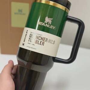 May include: A green and black Stanley Quencher H2.0 tumbler with a black handle. The tumbler has a gold band and the Stanley logo. The text on the tumbler reads "QUENCHER H2.0 TUMBLER" and "40 OZ | 1.18 L".