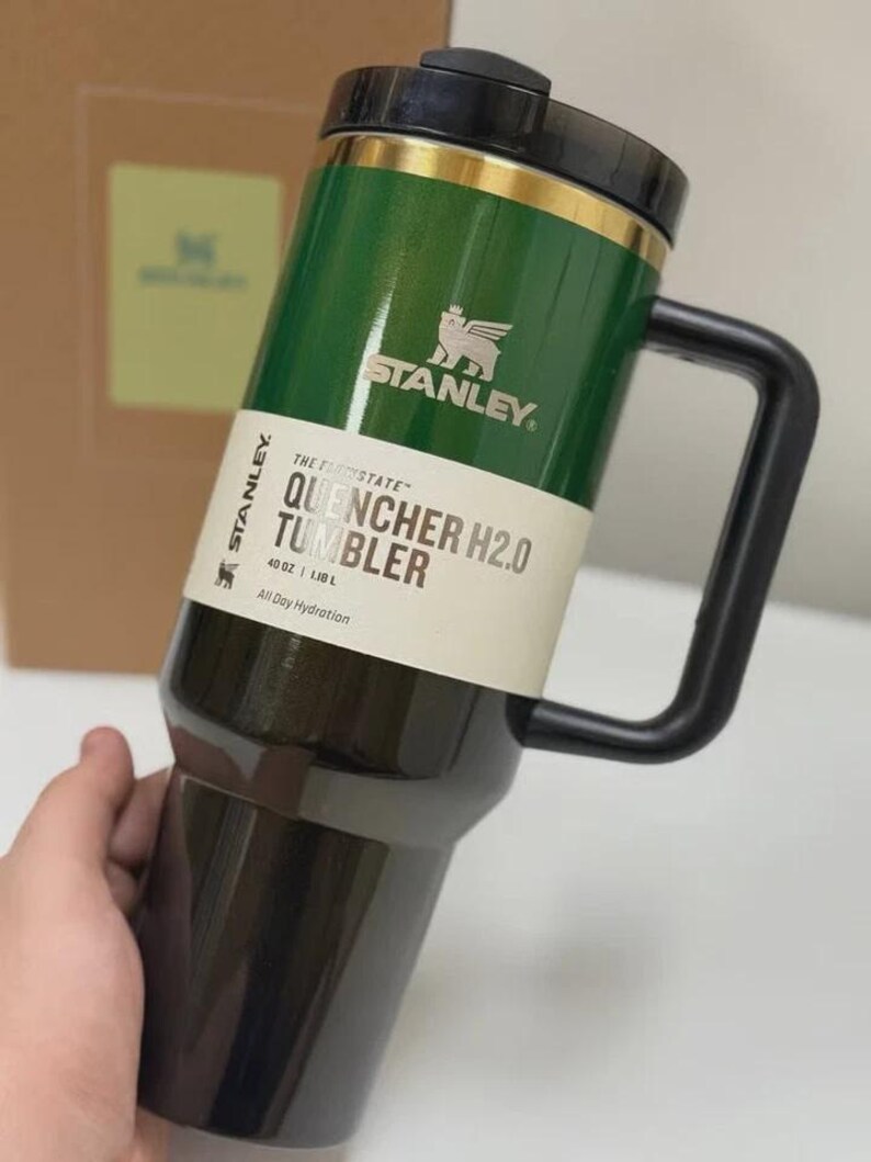 May include: A green and black Stanley Quencher H2.0 tumbler with a black handle. The tumbler has a gold band and the Stanley logo. The text on the tumbler reads "QUENCHER H2.0 TUMBLER" and "40 OZ | 1.18 L".