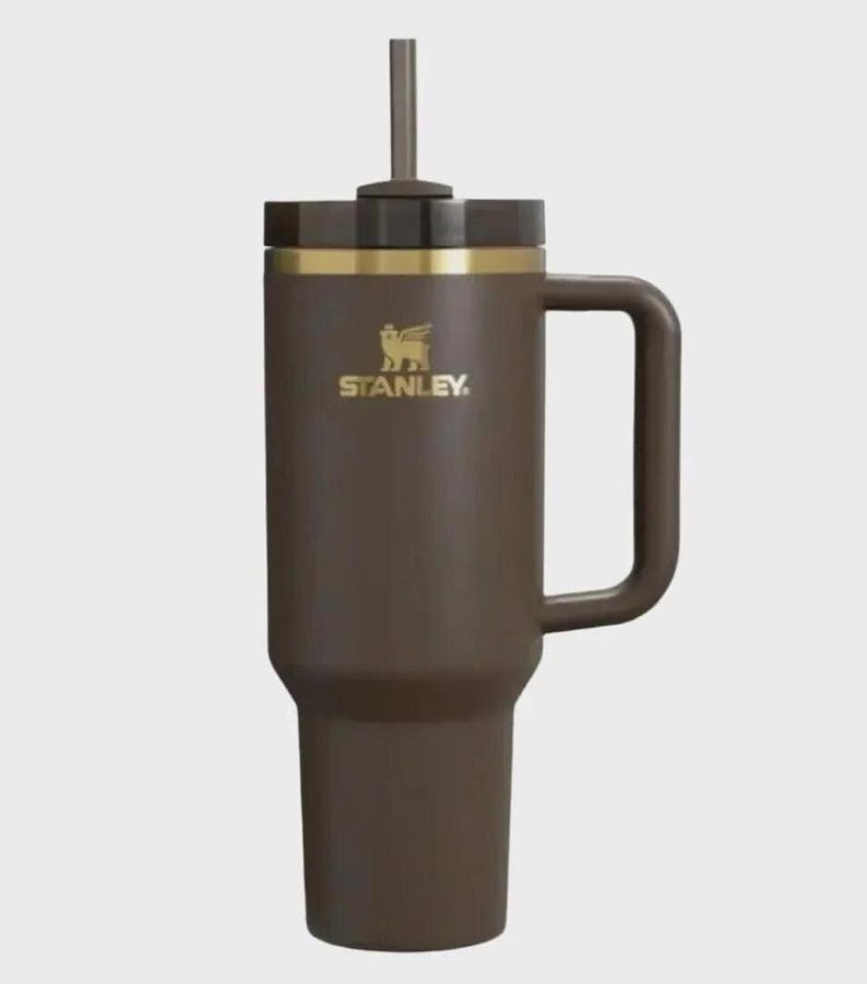 Puede incluir: Un vaso Stanley marr&oacute;n con asa y pajita. El vaso tiene una banda dorada y el logotipo de Stanley en dorado. Est&aacute; dise&ntilde;ado para mantener las bebidas calientes o fr&iacute;as durante horas. Capacidad aproximada de 1,2 litros.