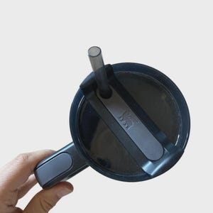 May include: A black travel mug lid with a gray handle and a black straw. The lid features a gray accent with a logo. The mug is designed for easy carrying and use.