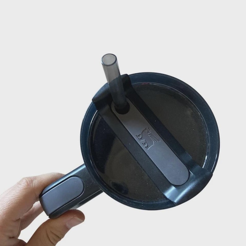 May include: A black travel mug lid with a gray handle and a black straw. The lid features a gray accent with a logo. The mug is designed for easy carrying and use.