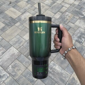 May include: Green Stanley Quencher H2.0 tumbler with a black handle and straw. The tumbler has a gold band and the Stanley logo. The tumbler is held by a person. The tumbler is 40 oz / 1.18L.