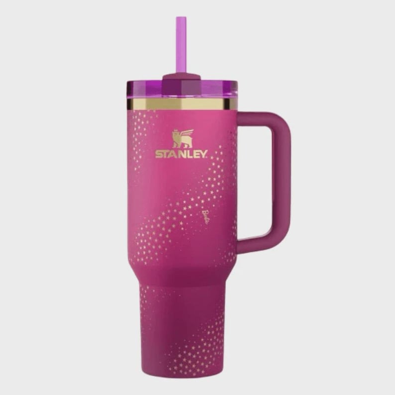 May include: A pink Stanley tumbler with a handle and straw. The tumbler has a gold rim and the Stanley logo. It features a pattern of gold stars and the word "Barbie" on the side.