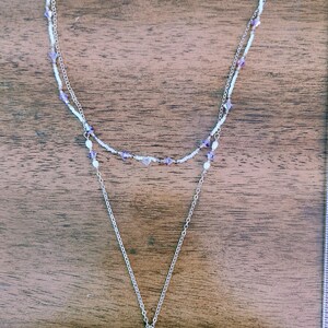 May include: A layered necklace with a silver chain and a pendant. The necklace features white and purple beads, with a teardrop-shaped crystal pendant. The background is a wooden surface.
