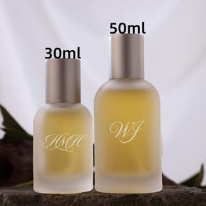 Könnte beinhalten: Zwei parfümflaschen aus mattiertem Glas, eine 30-ml- und eine 50-ml-Flasche, mit silbernen Verschlüssen. Die Flaschen enthalten eine gelbe Flüssigkeit und haben weiße Schriftinitialen. Der Hintergrund ist ein weiches Weiß mit einer Rose und dunklen Blättern.