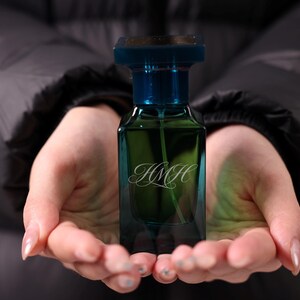 May include: A rectangular perfume bottle with a dark teal body and a square, dark blue cap. The bottle contains a light green liquid and the initials "HMH" are written in a cursive font on the front.