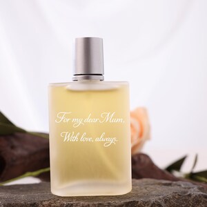 Peut inclure: Un flacon de parfum rectangulaire avec un bouchon argenté, rempli d'un liquide jaune clair. Le flacon porte l'inscription "For my dear Mum, With love, always." Le flacon est posé sur une surface en pierre, avec une branche en bois et des roses couleur pêche en arrière-plan.