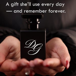 May include: A black rectangular perfume bottle with a black cap, held in open hands. The bottle has the white letters "DG" in a cursive font. The image includes the text: "A gift she'll use every day — and remember forever."
