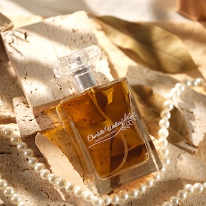 Puede incluir: Un frasco de perfume rectangular con una tapa transparente facetada y un cuello plateado. El líquido ámbar es visible en el interior. La botella está decorada con el texto "Charlotte Walker 22nd December 2025" y un diseño floral. Un collar de perlas y hojas secas están en el fondo.
