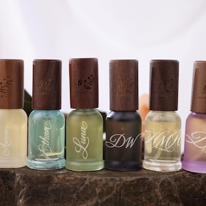 May include: Six perfume bottles with wooden caps are arranged on a stone surface. The bottles contain different colored liquids, including yellow, blue, green, and purple. Each bottle has a name or initials printed on it.