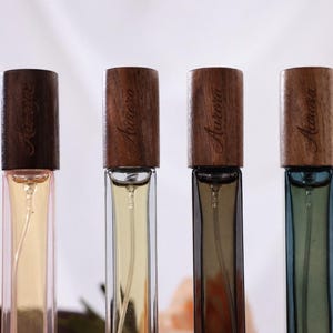 以下が含まれることがあります： 木製のキャップが付いた4つの香水瓶で、それぞれに「Aurora」と書かれています。ボトルには、薄い黄色、透明、茶色、青色の異なる色の液体が入っています。ボトルは白い背景に表示されています。