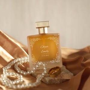 Puede incluir: Un frasco de perfume ámbar cuadrado con tapa dorada, adornado con diseños florales blancos y los nombres "Oliver & Emily" y la fecha "12.12.2015". Un collar de perlas y un anillo dorado descansan cerca del frasco sobre una superficie beige, con un fondo de tela dorada.