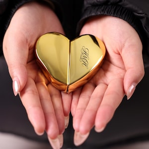 Puede incluir: Un estuche compacto dorado en forma de corazón con las iniciales "M&A" grabadas en la parte delantera. El estuche compacto se sostiene en manos abiertas sobre un fondo oscuro. El estuche compacto es de color dorado brillante.
