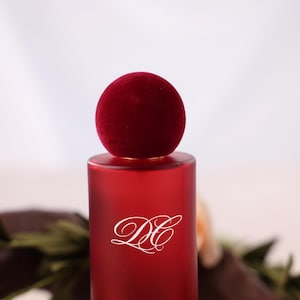 May include: A red cylindrical perfume bottle with a matching velvet-covered stopper. The bottle has a white script logo. The background is a soft white with floral accents.