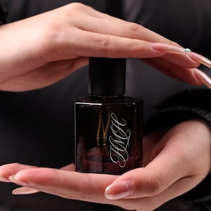 Peut inclure: Un flacon de parfum rectangulaire marron foncé avec un bouchon noir. Le flacon porte les initiales "HMH" en écriture cursive. Le flacon est tenu dans deux mains sur un fond sombre.