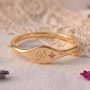 May include: A gold signet ring featuring an engraved cursive letter D and a small star-shaped accent set with a tiny pink gemstone. The ring has a polished finish and a smooth, rounded band, showcasing a delicate and refined design.
