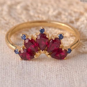 May include: A gold ring featuring a curved arrangement of five pear-cut red rubies and four small round blue sapphires. The gemstones are set in a yellow gold band. The text BespokeBlingAtelier is visible in the corners of the image.