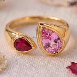 May include: A gold bypass ring featuring two pear-cut gemstones set in polished metal. One stone is a deep red, while the larger stone is a light pink. The band is smooth and open-ended, showcasing a contemporary design with clear, faceted stones.