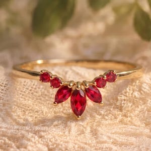 Pode incluir: Um anel de ouro com uma série de pedras preciosas vermelhas vibrantes. O design do anel apresenta uma disposição curva de pedras de corte marquesa, criando uma estética delicada e elegante. O anel está sobre um fundo macio e texturizado.