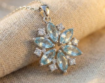 Puede incluir: Un collar de plata con un colgante en forma de flor. El colgante presenta piedras preciosas de color azul claro dispuestas en un patrón floral, con piedras transparentes más pequeñas entre los pétalos. El collar está sobre una tela beige.