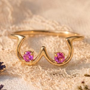 May include: A gold ring with a unique design, featuring two pink gemstones. The ring's shape is artistic and eye-catching, set against a soft, textured background with floral accents. The gemstones are round and vibrant.